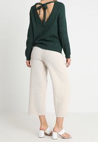 Grön stickad tröja med djup V-rygg och knytdetalj, tillsammans med krämfärgade vida byxor och vita klacksandaler med blockklack.