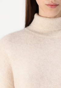 Gros plan sur une personne portant un pull à col roulé doux de couleur crème, avec de longs cheveux bruns visibles sur un fond blanc.