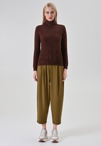 Maglione marrone a collo alto con finitura texturizzata, abbinato a pantaloni plissettati verde oliva e sneaker sportive grigio chiaro.