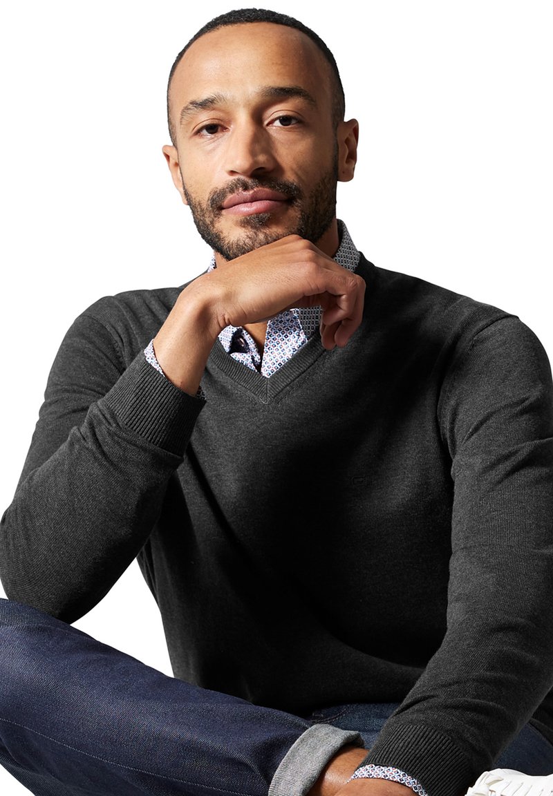 Maglione grigio scuro a V realizzato in tessuto morbido, indossato sopra una camicia a righe blu e bianca, abbinato a jeans blu e sneakers bianche.