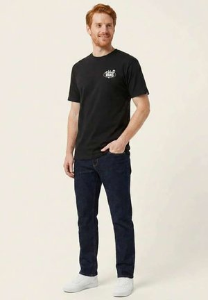 Homme roux souriant, portant un t-shirt Vans noir, un jean bleu foncé et des baskets blanches, debout avec une main dans la poche.