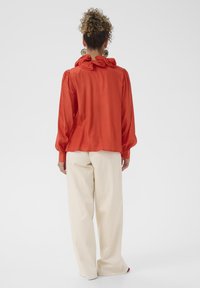 Blouse en satin rouge avec manches bouffantes et col festonné, associée à un pantalon large crème. Texture lisse, design simple sans motifs.
