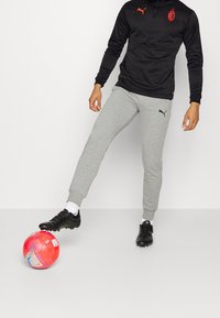 Maglietta sportiva nera a maniche lunghe con logo e dettagli rossi, abbinata a pantaloni della tuta grigi a gamba stretta e scarpini da calcio neri, posizionati vicino a un pallone da calcio rosa.