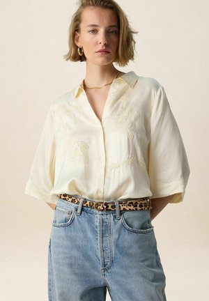Jonge vrouw in een licht crèmekleurige blouse met borduursels en ellebooglange mouwen, een riem met luipaardprint en een high-waisted blauwe spijkerbroek tegen een effen achtergrond.