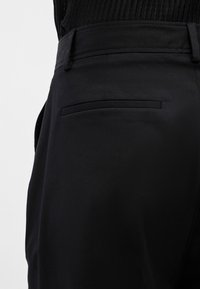 Tuvplānā redzama persona, kas valkā melnas piegrieztas bikses ar aizmugurējo šuvju kabatu un melnu ribas tekstūras augšu.