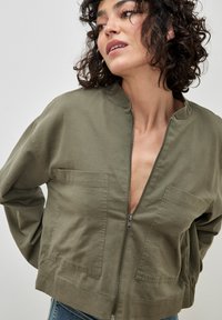 Olive green bomber jacket with a zip front, two chest pockets, and a cropped fit. Made of soft fabric with a smooth texture.