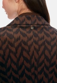 Brown patterned blazer with a black chevron design. Features a notched collar and a round metal logo accent. Smooth texture, tailored fit.