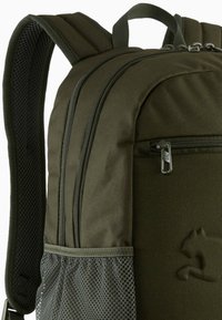 Sac à dos vert olive doté de bretelles rembourrées réglables, de multiples compartiments zippés et d'une poche latérale en mesh. Détails en tissu texturé visibles.