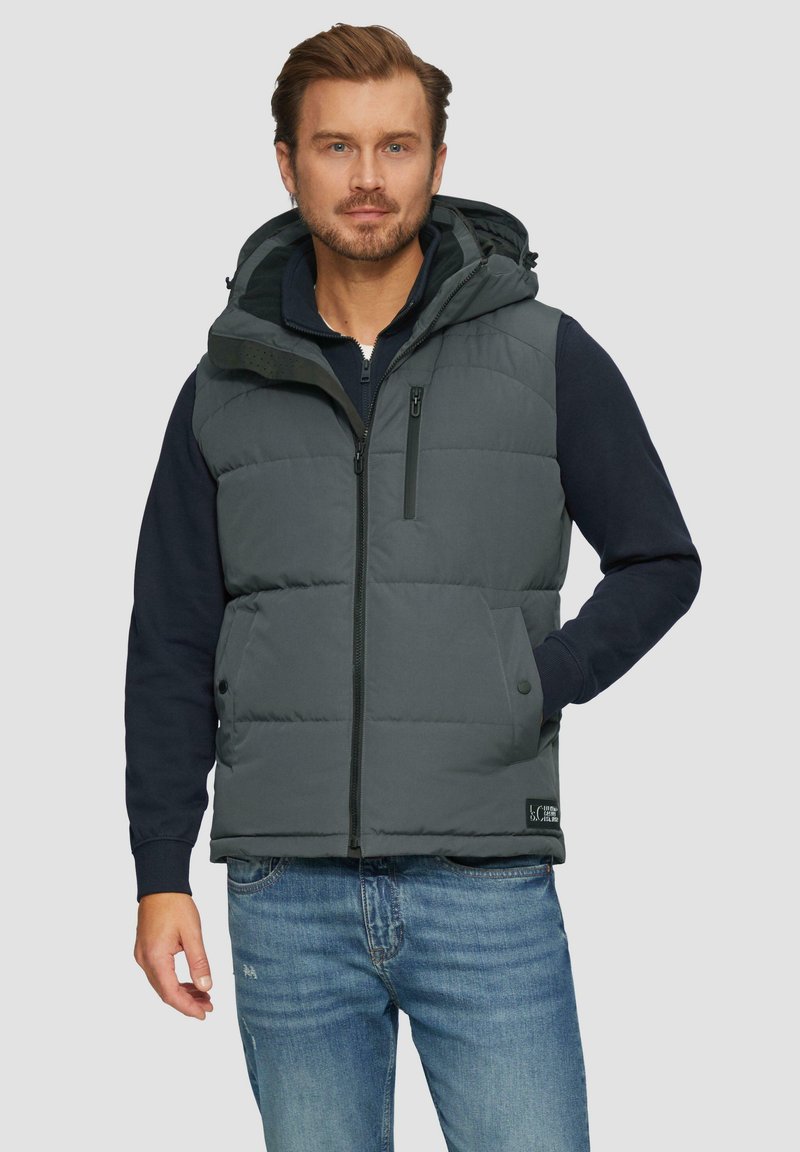 Graue Steppweste mit Kapuze, gestepptem Design, Reißverschluss vorne, seitlichen Taschen und einem Logo-Patch am Saum. Gestylt über einem dunkelblauen Sweater.