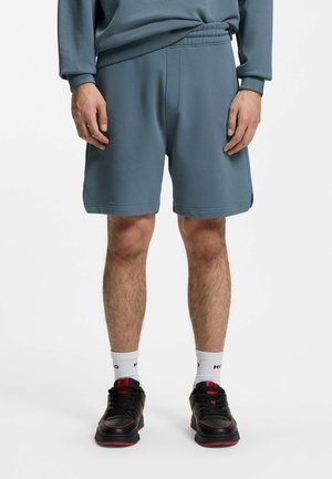 Mand iført blå elastiske shorts med elastik i taljen, hvide crew-sokker med tekst og sorte sneakers med røde detaljer, stående på en hvid baggrund.