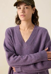 Jeune femme aux yeux verts portant une casquette à imprimé léopard, des boucles d'oreilles en or, et un pull V en col violet, les bras croisés.