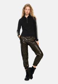 Schwarzer Reißverschluss-Pullover mit geripptem Design und weißen Streifen an den Ärmeln, kombiniert mit dunkelgrünen, gemusterten Leder-Jogginghosen und schwarzen Stiefeln.