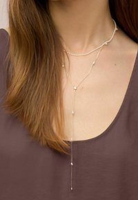 Femme aux longs cheveux bruns portant un haut violet et deux colliers superposés : un collier de perles court et une longue chaîne dorée avec de petites perles.