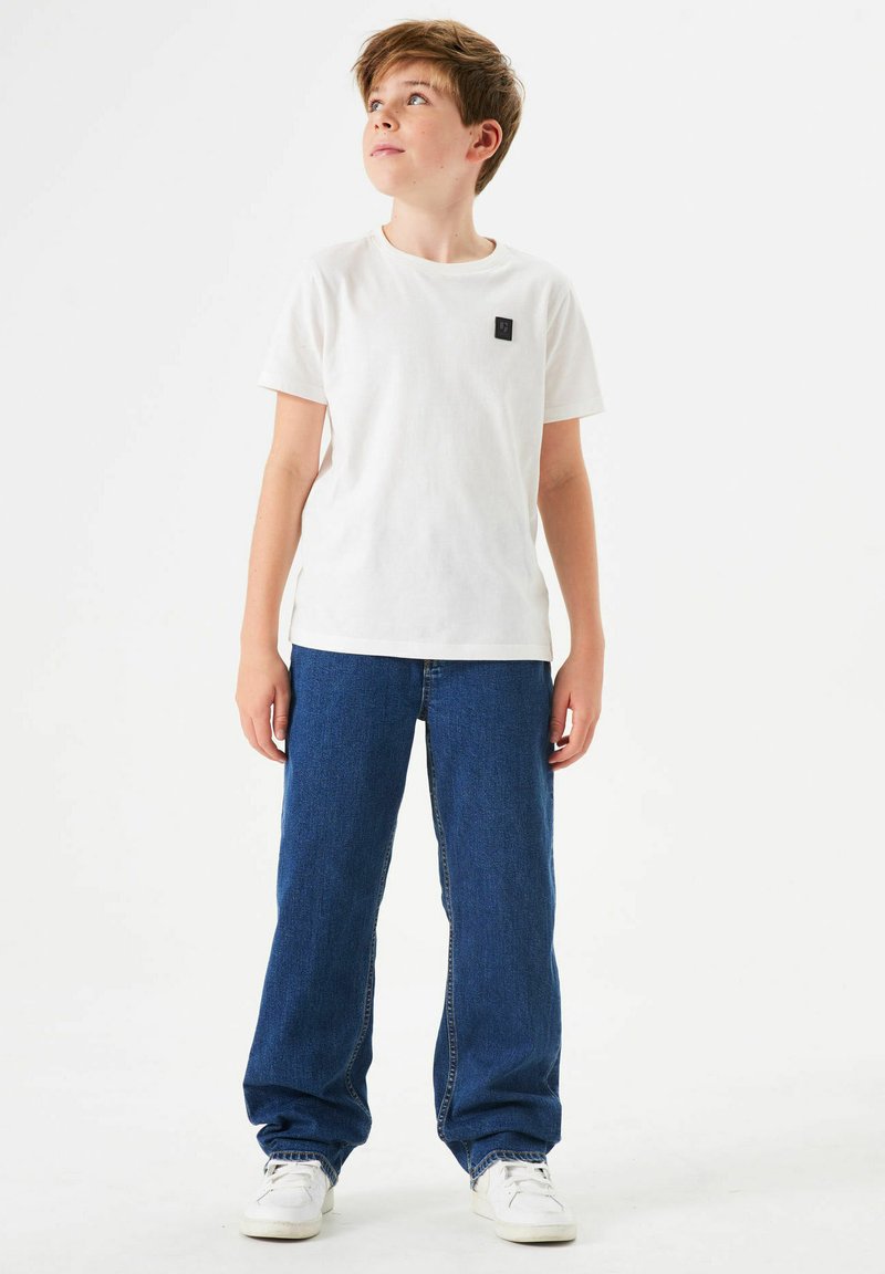 Camiseta blanca de algodón con un parche de logo negro, combinada con jeans de denim azul sueltos y zapatillas blancas, de pie frente a un fondo sencillo.