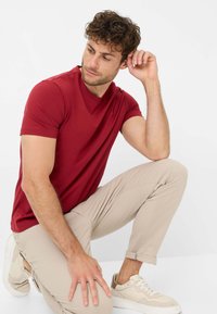 Rotes Kurzarm-T-Shirt mit rundem Halsausschnitt, kombiniert mit hellbeigen, schlanken Hosen und beigenSneakern. Strukturierter Stoff, lässige Passform.