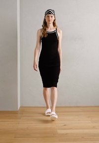 Abito nero senza maniche con bordi bianchi e design aderente, abbinato a un foulard a righe bianche e nere e zoccoli bianchi, su uno sfondo grigio.