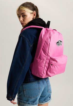 Joven con chaqueta de mezclilla y pantalones cortos, llevando una mochila Vans de color rosa brillante en la espalda, mirando por encima del hombro.