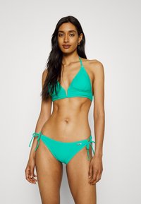 Turkio spalvos bikini maudymosi kostiumėlis su trikampiu viršumi ir reguliuojamomis juosmens šoninėmis juostelėmis. Lygus tekstūras, minimalus dizainas ir mažas metalinis logotipas ant juosmens.