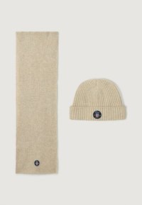 Bufanda de punto acanalado beige y gorro a juego, ambos con un parche de logo circular con acentos. Textura suave y diseño sencillo.