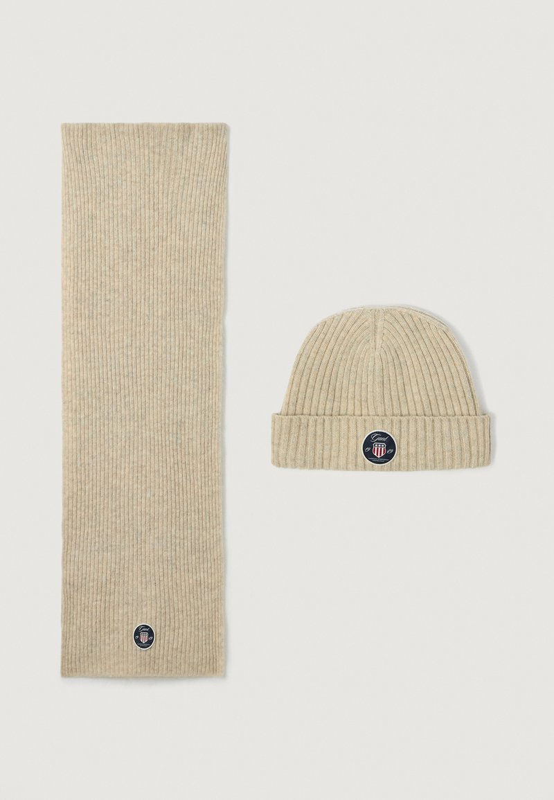 Bufanda de punto acanalado beige y gorro a juego, ambos con un parche de logo circular con acentos. Textura suave y diseño sencillo.