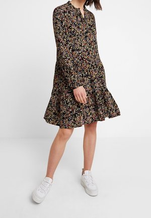 Woman wearing a black long-sleeve floral dress and white sneakers, standing with legs apart against a plain background.