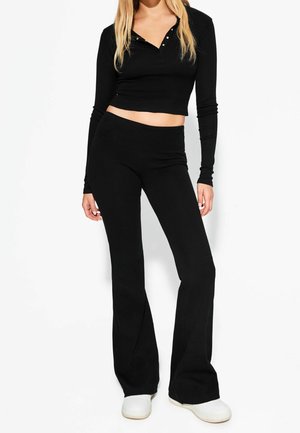 Frau trägt ein schwarzes langärmeliges Crop-Top und hoch taillierte schwarze ausgestellte Hose mit weißen Slip-On-Schuhen vor weißem Hintergrund.