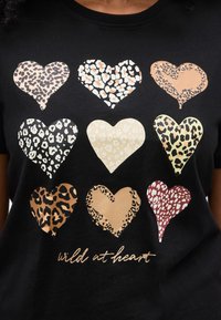 Εννέα καρδιές με σχέδιο λεοπάρδαλης σε διάφορα χρώματα, διατεταγμένες σε πλέγμα σε μια μαύρη μπλούζα με το κείμενο «wild at heart» από κάτω.