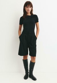 Sort tætsiddende top med korte ærmer, kombineret med højtaljede sorte plisserede shorts. Sorte knæhøje støvler fuldender outfittet.