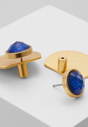 Goldene Ohrringe mit facettierten runden blauen Edelsteinen, ein Paar auf einer flachen halbrunden Basis, auf einer weißen Oberfläche präsentiert.
