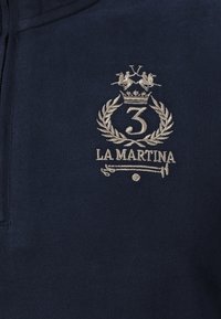 Tmavě modrá látka s béžovým vyšitým logem zobrazujícím korunované číslo 3, vavřínový věnec, překřížené pálky na pólo a text "LA MARTINA".
