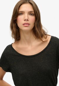 Camiseta de manga corta en gris oscuro con un escote amplio, que presenta sutiles motas amarillas y un pequeño logo en el pecho. Tejido suave y con textura.