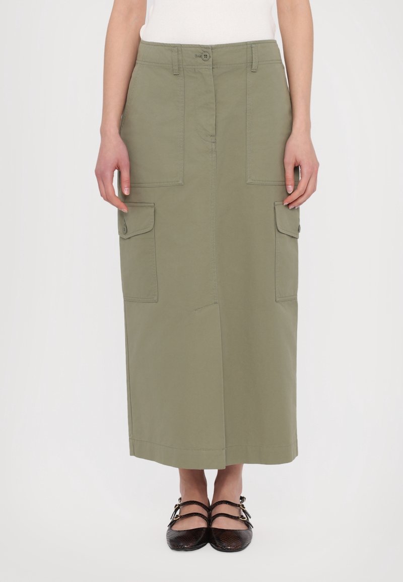 Jupe midi vert olive avec boutons sur le devant et poches à rabat, portée avec des chaussures plates noires à double bride et un haut blanc.