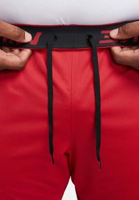 Mani che stringono i lacci neri dei pantaloni sportivi rossi con vita nera e scritte rosse.