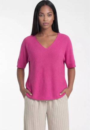 Kobieta z prostymi czarnymi włosami nosi jasnoróżowy sweter z krótkim rękawem i dekoltem w serek oraz beżowe spodnie w cienkie paski, pozując z rękami w kieszeniach.