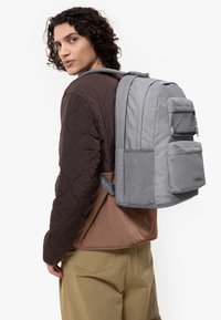 Mochila de tela gris con múltiples bolsillos, forma rectangular y acentos de malla. Llevada sobre una chaqueta marrón acolchada y pantalones beige.