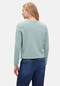 Jasnozielony sweatshirt z długimi rękawami, luźnym krojem, okrągłym dekoltem i skróconą długością. Noszony z niebieskimi dżinsami.