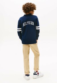 Navyblaues Langarm-Polohemd mit weißem "HILFIGER"-Text und Streifen an den Ärmeln, beigen Hosen und weißen Sneakern mit navyblauen und roten Akzenten.