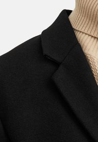 Schwarzer Wollmantel mit Reverskragen und glatter Textur, über einem ribbenförmigen beigen Pullover getragen. Sichtbare Nähte verleihen dezente Details.