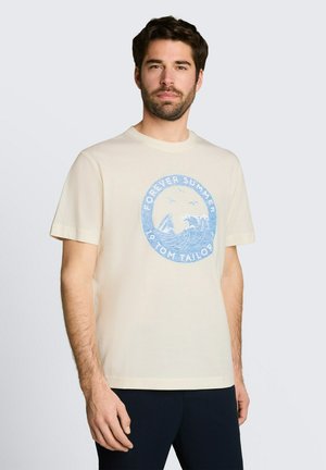 Mężczyzna noszący beżową koszulkę z niebieskim okrągłym wzorem "Forever Summer" z falą, żaglówką i mewami, stojący na tle prostego jasnego tła.