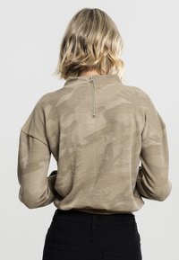 Beige camouflert mønsteret genser med høy hals, halv glidelås bak og nedfelte ermer. Myk ribbestrikket tekstur.