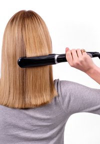 Un lisseur à cheveux noir tenu dans une main, avec une finition polie. Les cheveux sont raides, blonds et de longueur au niveau des épaules sur un fond blanc.