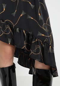 Mustat epäsymmetriset mekko, jossa on röyhelöpohja, koristeltu kultaisilla ja vihreillä tupsukuviolla. Mustat kiiltävää nahkaa polvipituiset saappaat näkyvissä.