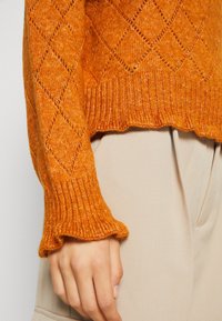 Pomarańczowy sweter dzianinowy z wzorem w romby, prążkowanymi mankietami z falbanką i teksturowanym wykończeniem na beżowej tkaninie.