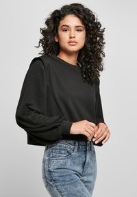 Femme avec des cheveux noirs bouclés portant un haut noir à manches longues bouffantes et un jean bleu clair taille haute, se tenant devant un fond gris.