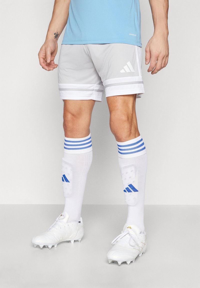 Athlète masculin portant un short Adidas blanc et gris, des chaussettes blanches à rayures bleues, des protège-tibias et des chaussures de football blanches, debout sur un fond blanc.