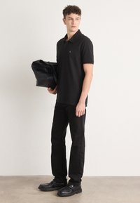 Zwarte polo met een groen logo, gecombineerd met zwarte broek en zwarte schoenen. De persoon houdt een zwarte leren tas vast.