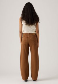 Vrouw met lang krullend haar, gekleed in een witte zonder mouwen gehaakte top en hoge taille brede bruine broek, staand tegen een effen lichte achtergrond.