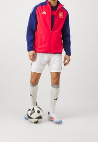 Red and navy athletic jacket with zipper, Spain logo on chest, paired with white shorts, knee-high socks, and black-and-white soccer ball.