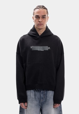 Persona con trenzas lleva una sudadera negra con texto blanco desvaído y vaqueros azul claro, desgastados y sueltos, sobre un fondo liso y claro.