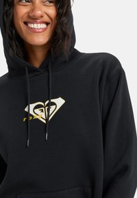 Sudadera con capucha negra que presenta un gran logotipo en forma de corazón en blanco y dorado. Textura suave con un bolsillo canguro y cordones ajustables.